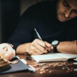 Crop elegant man taking notes in journal while working at desk with coins and piggybank in lamplight