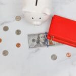 woman writing in notebook next to piggy bank and stack of coins, emergency savings guide on desk